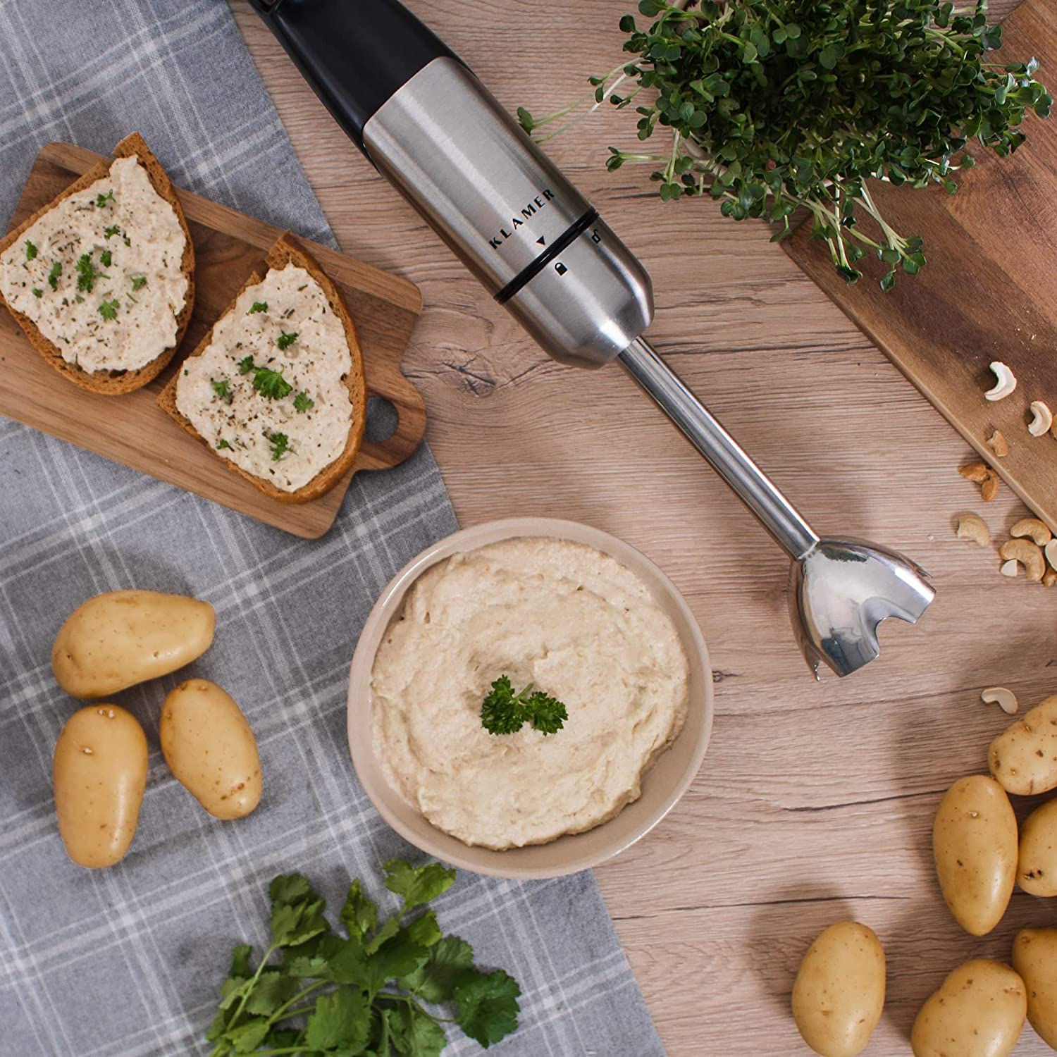 Lebensmittelzubereitung: Hummus, Brot, Kartoffeln, Kräuter und ein Mixer auf Holz. Hummus in Schüssel, belegte Brotscheiben.