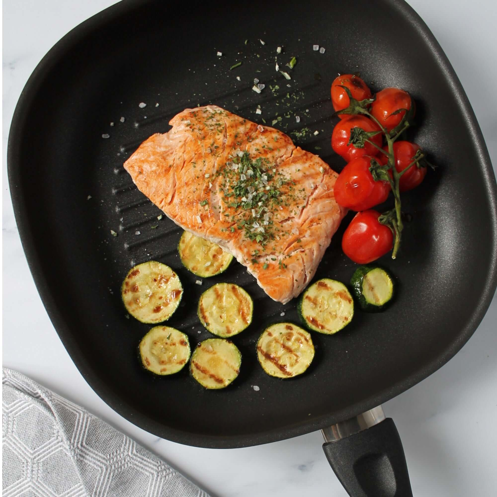 Eine Pfanne mit gekochtem Lachs, gegrillten Zucchinischeiben und Tomaten. Das Essen ist garniert.