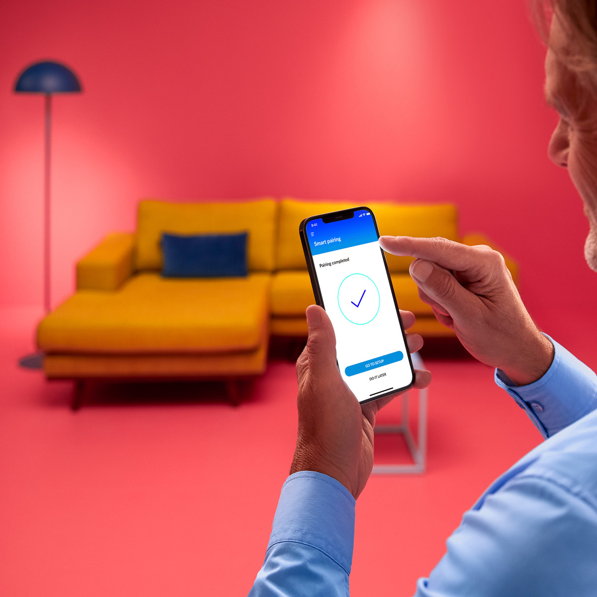 Person hält Telefon mit 'Pairing abgeschlossen' und Haken. Gelbe Couch und rosa Wand im Hintergrund.