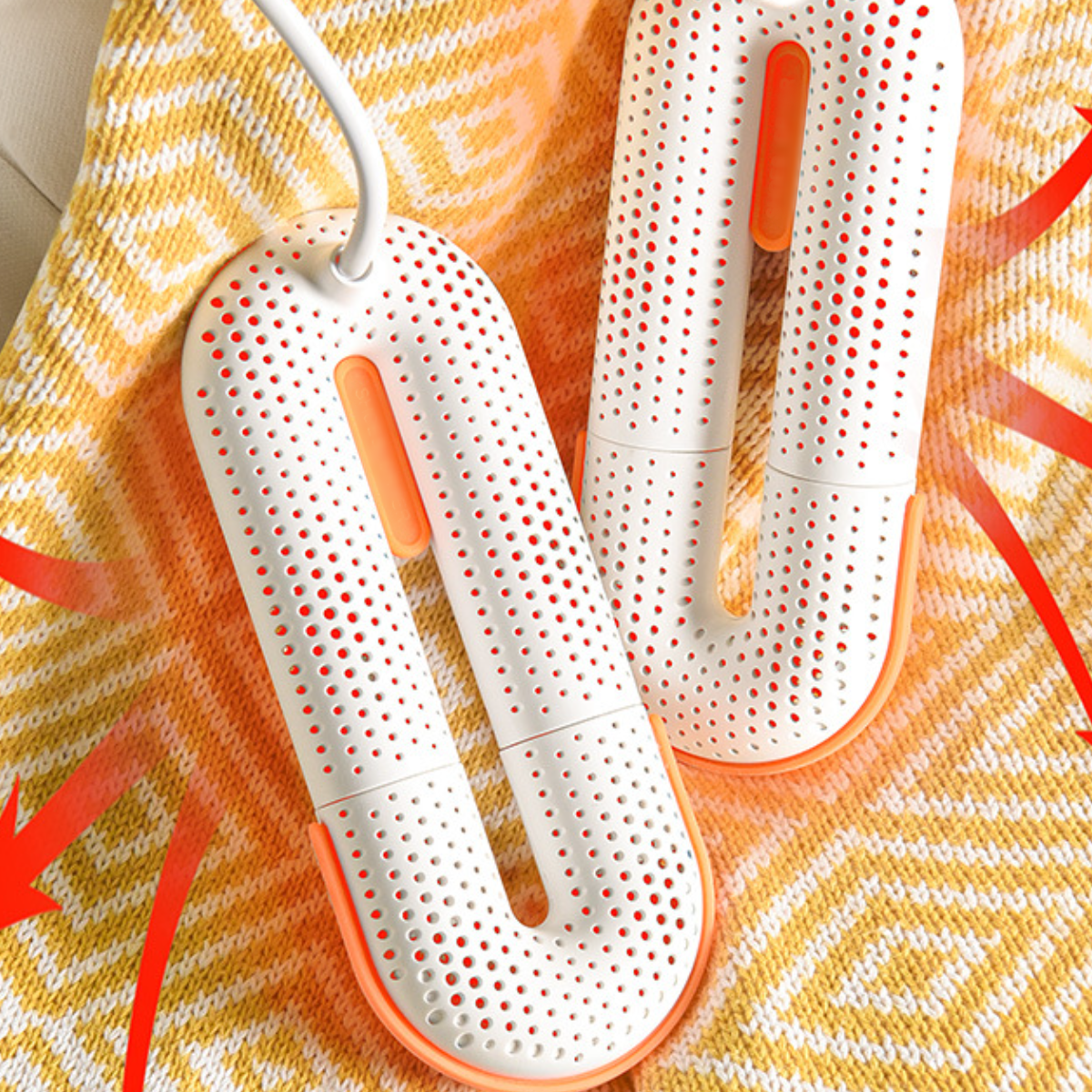 Zwei weiße Schuhtrockner mit orangefarbenen Akzenten auf einer gelben, gemusterten Oberfläche. Rote Pfeile zeigen den Luftstrom an.