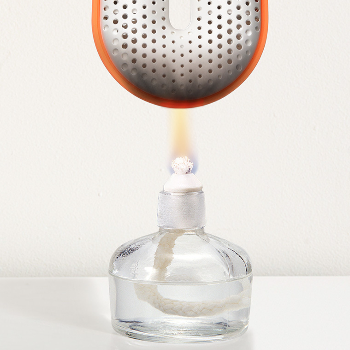 Klare Glasflasche mit Docht und Flamme sowie einem dekorativen orangefarbenen Deckel. Weißer Hintergrund.