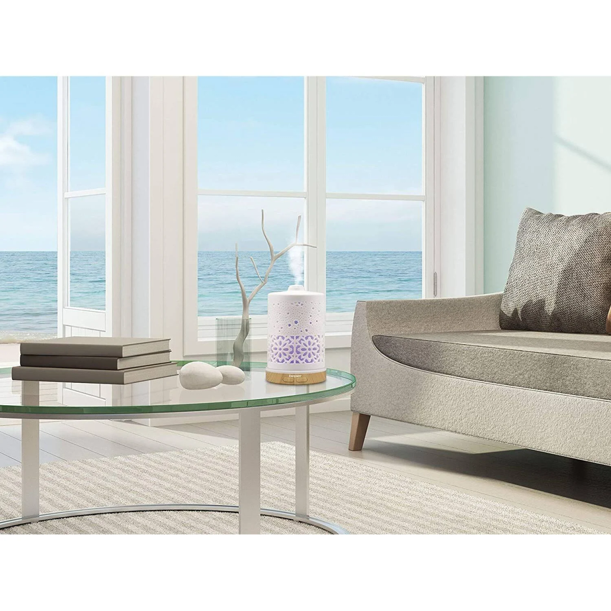 Ein ätherischer Öl-Diffusor auf einem Glastisch in der Nähe eines Fensters mit Meerblick. Eine Couch ist im Bild.