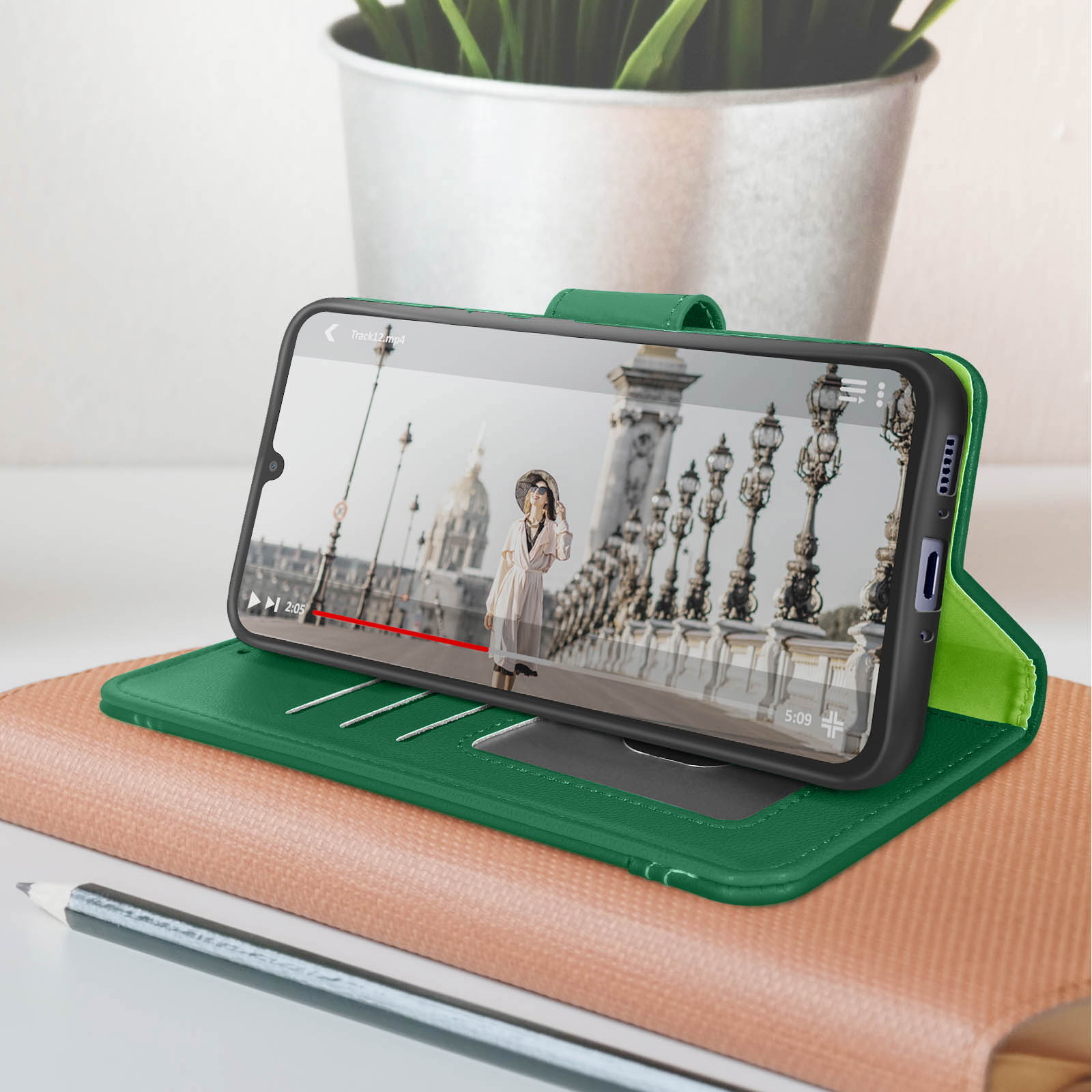 Custodia verde per telefono con un telefono all'interno, che riproduce video. Il telefono è su una scrivania, con una pianta.