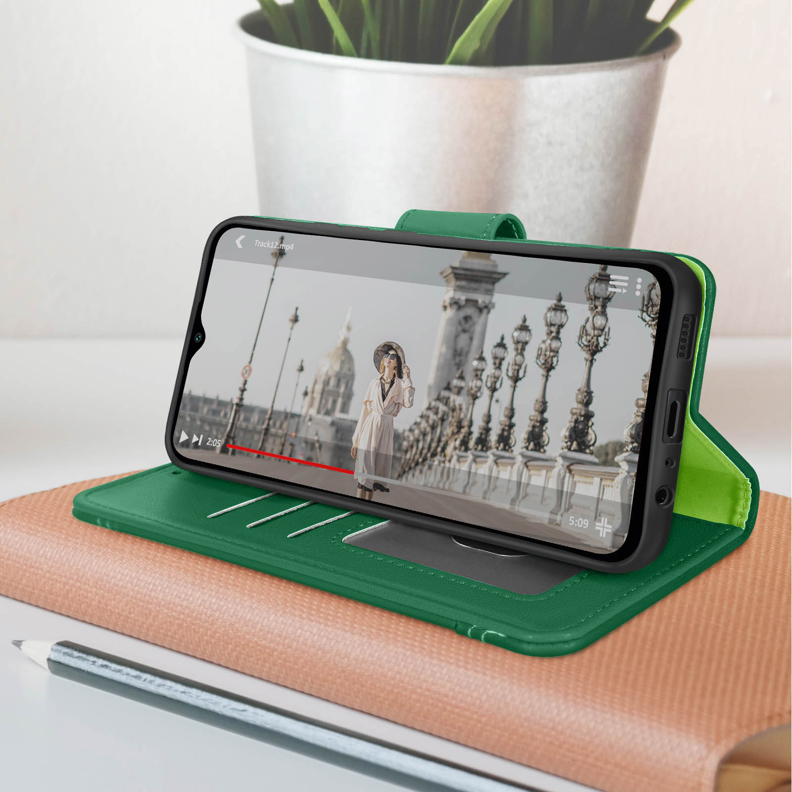 Un telefono in una custodia verde mostra un video di una donna su un ponte. Un taccuino e una penna sul tavolo.