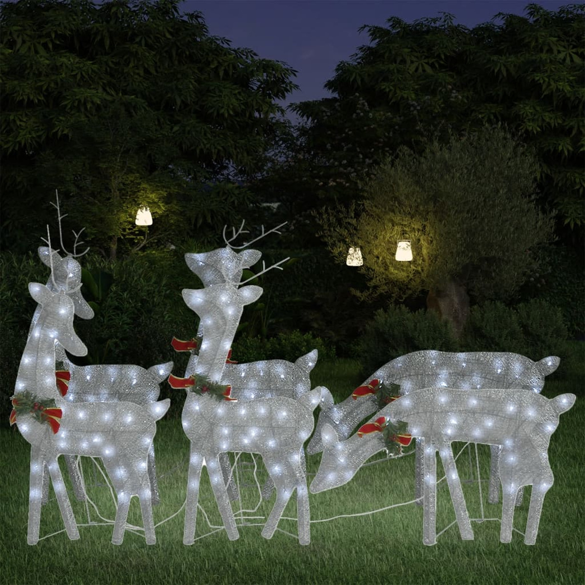 Beleuchtete Rentier-Anzeige im Garten bei Nacht. Bäume und Laternen im Hintergrund.