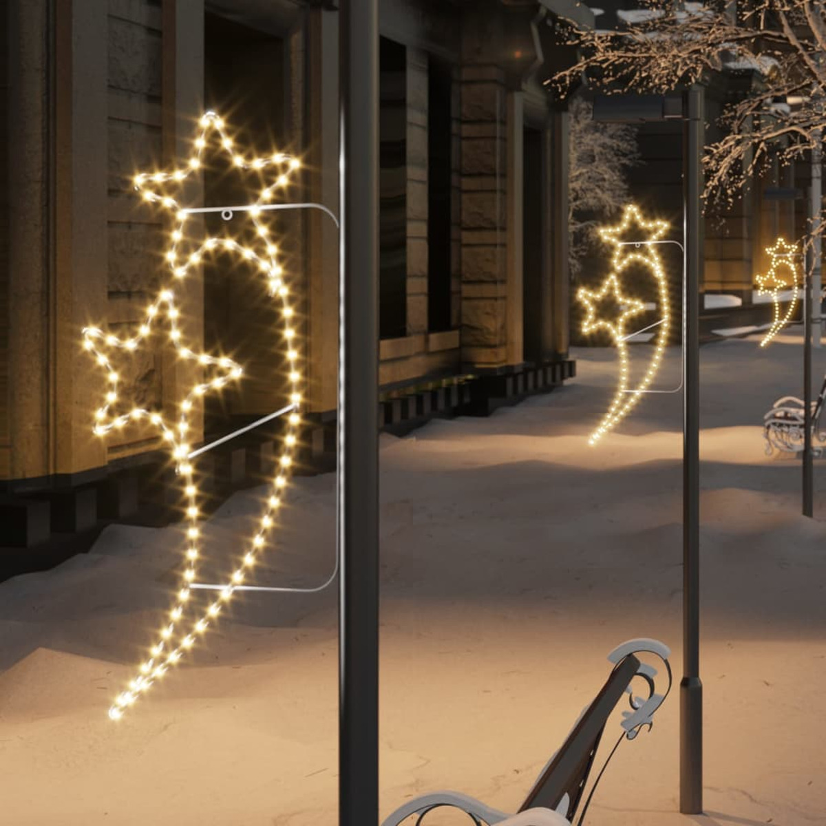 Weihnachtsbeleuchtung in Sternform an Laternenpfählen in einer verschneiten Straße bei Nacht. Eine Bank im Hintergrund.