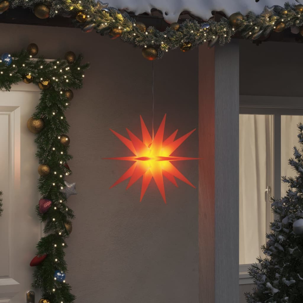 Ein großer, orangefarbener sternförmiger Weihnachtsschmuck hängt draußen an einem Haus, mit Schnee und Girlande.
