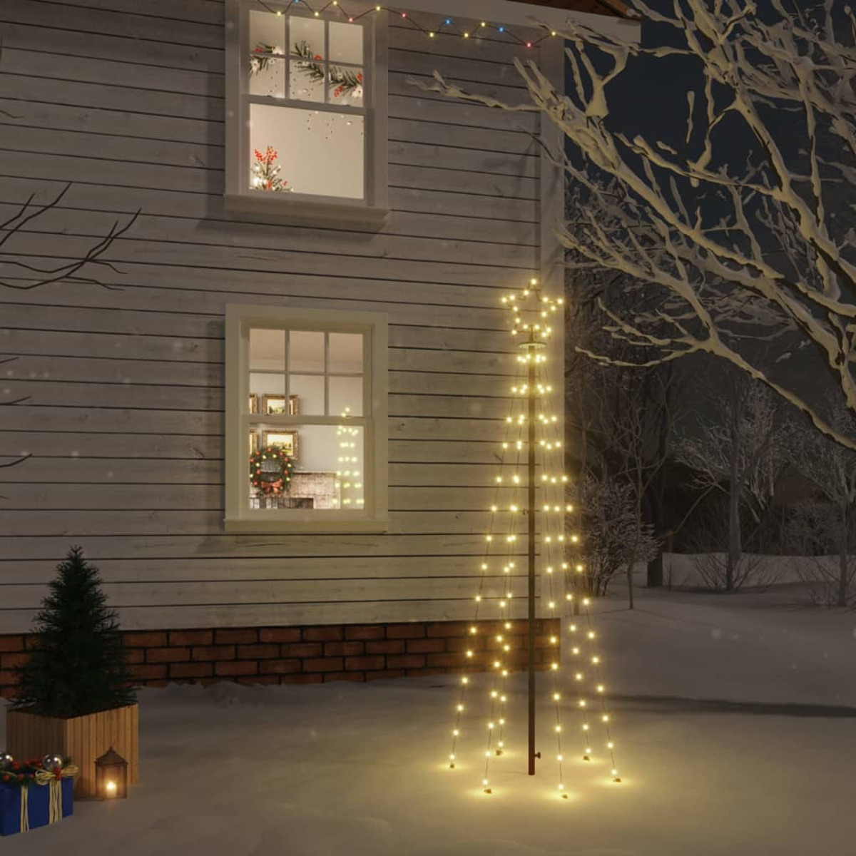 Ein Haus mit beleuchteten Fenstern und einem beleuchteten Baum im Schnee. Ein kleiner Baum und Geschenke sind davor. Nachtszene.