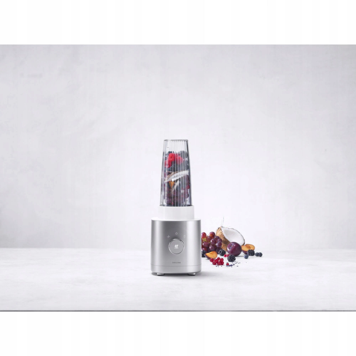 Blender ze składnikami. Ma szklany pojemnik z owocami i srebrną podstawę.