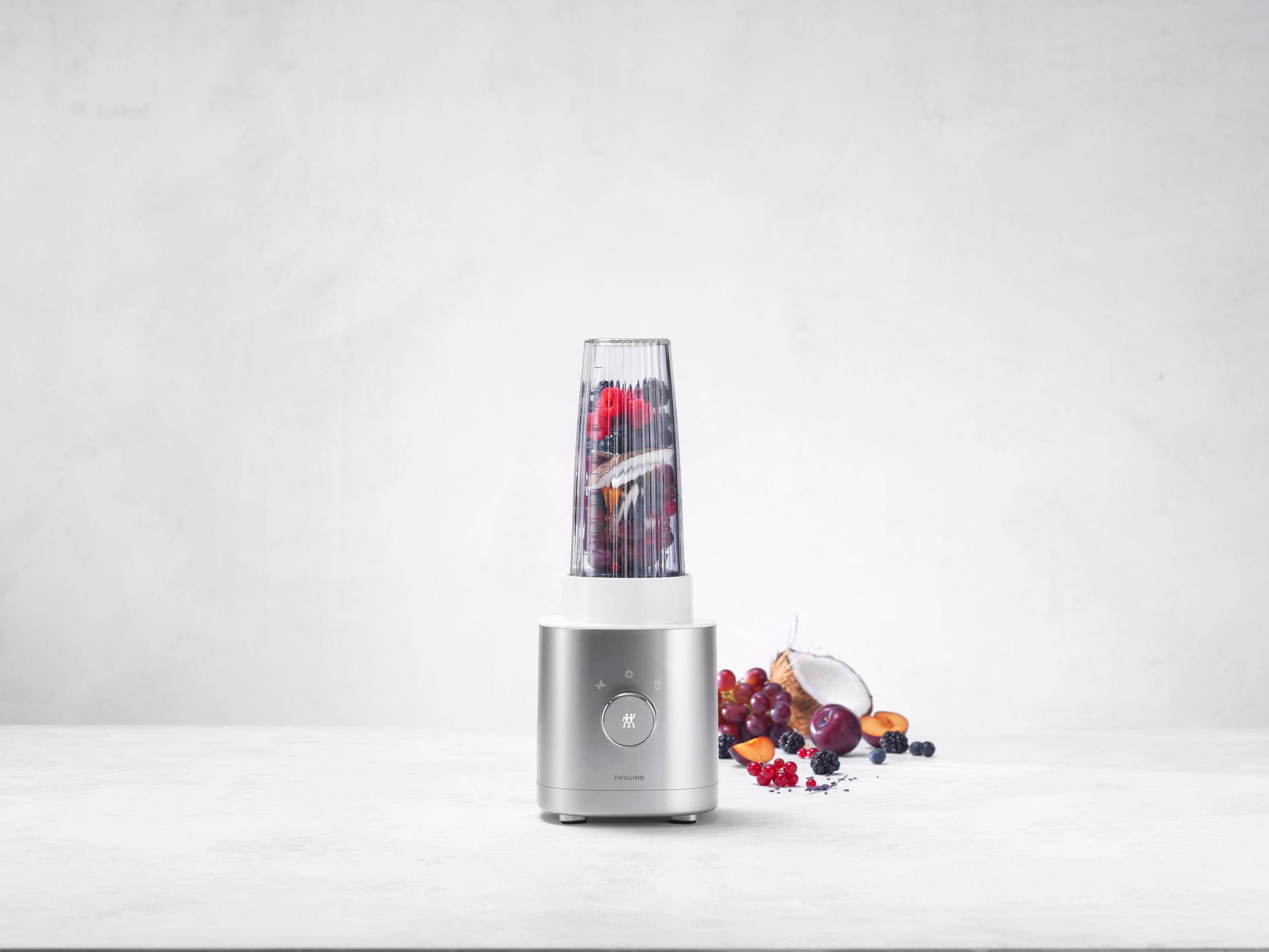 Srebrny blender z owocami. Białe tło. Owoce i kokos wokół blendera.