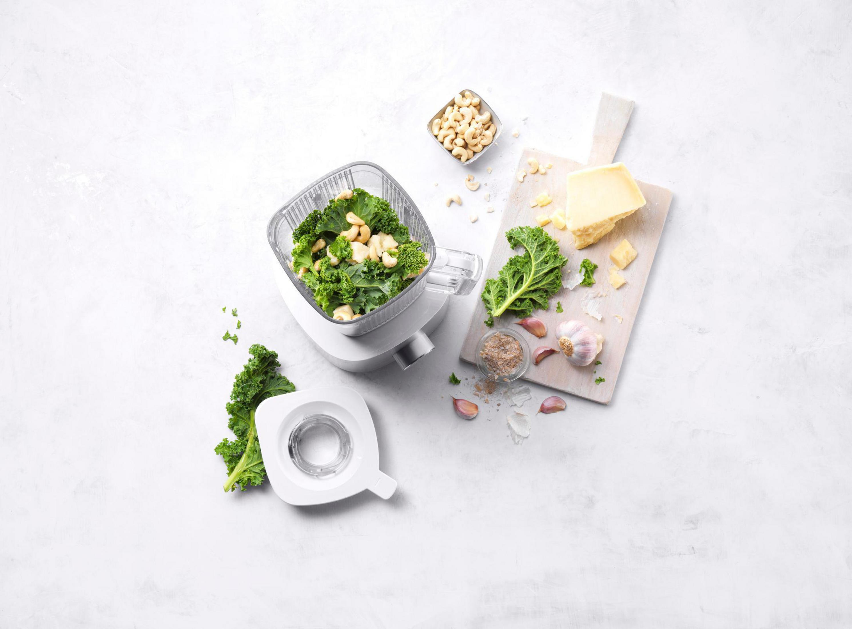 Blender z jarmużem, orzechami nerkowca. Ser i czosnek na desce. Białe tło.
