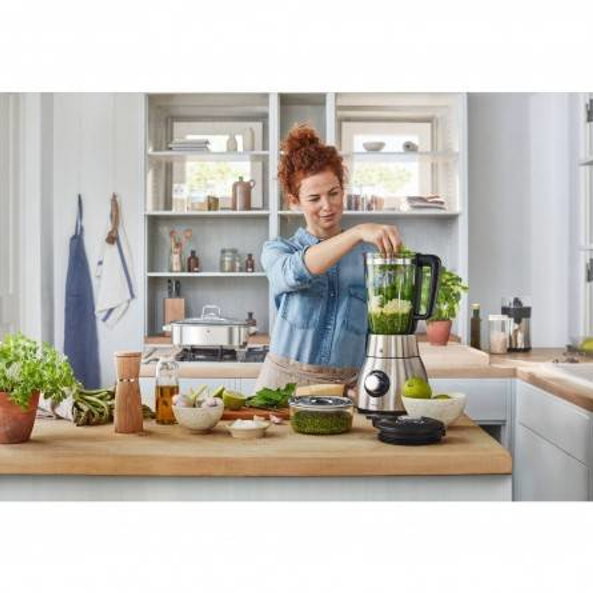 Kobieta przygotowująca jedzenie blenderem w kuchni. Różne składniki i przybory na blacie.