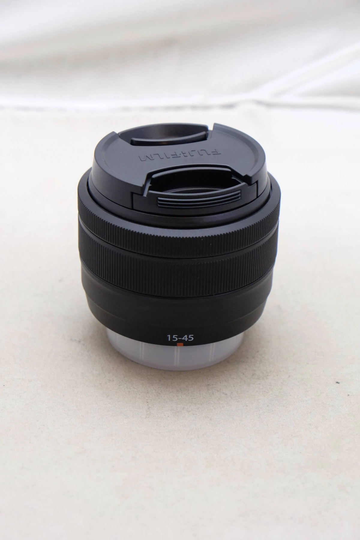 Czarny obiektyw aparatu oznaczony 15-45. Obiektyw ma górną pokrywę i leży na jasnej powierzchni.