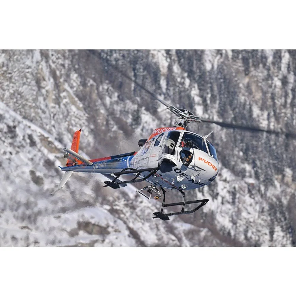 Un helicóptero gris y blanco volando frente a una cordillera cubierta de nieve.