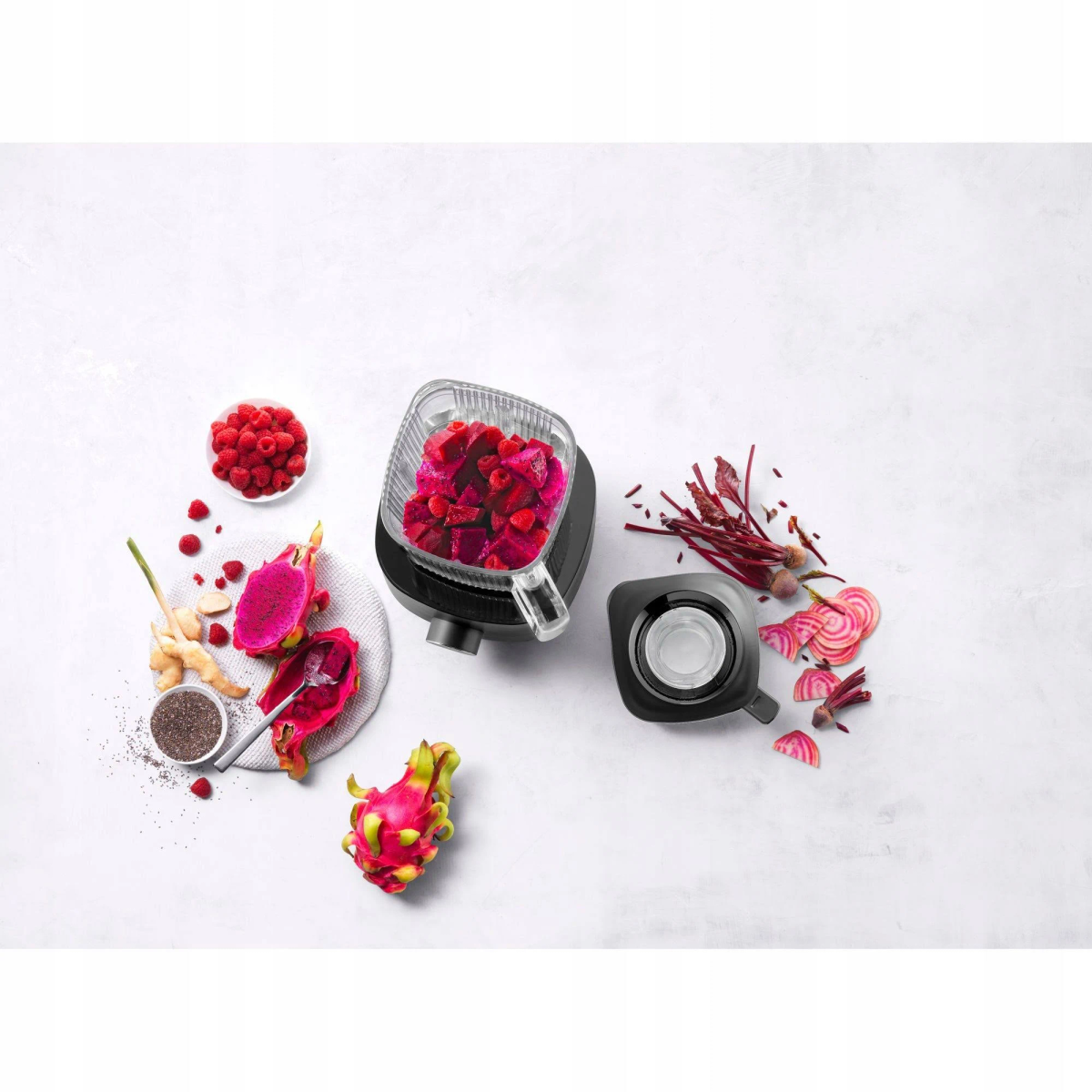 Blender jest otoczony składnikami takimi jak maliny, buraki i smoczy owoc na białej powierzchni.