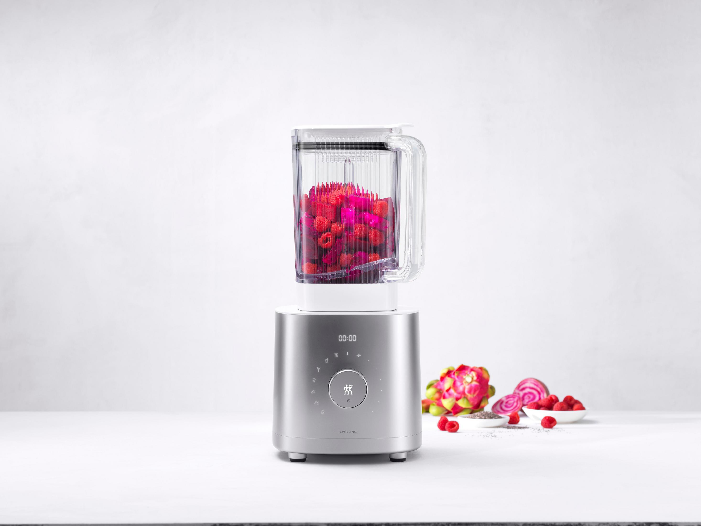 Blender z malinami w środku, na srebrnej podstawie. Różowe owoce i białe tło.