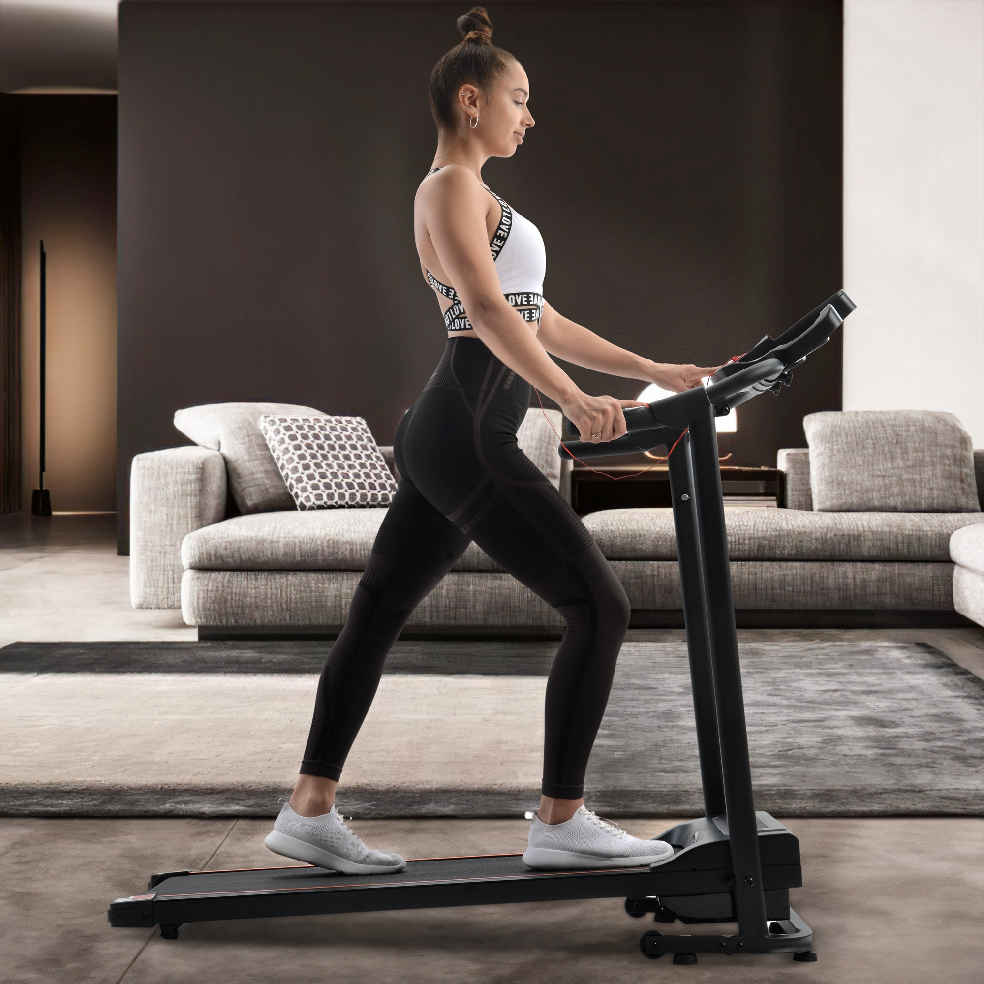 Frau auf dem Laufband im Wohnzimmer. Sie trägt ein weißes Oberteil, schwarze Leggings und weiße Schuhe.