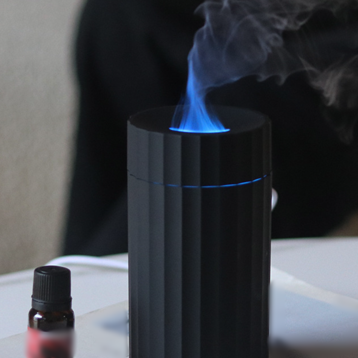 Ein schwarzer Diffusor gibt blauen Dampf ab. Daneben steht eine kleine Flasche, mit verschwommenem Hintergrund.