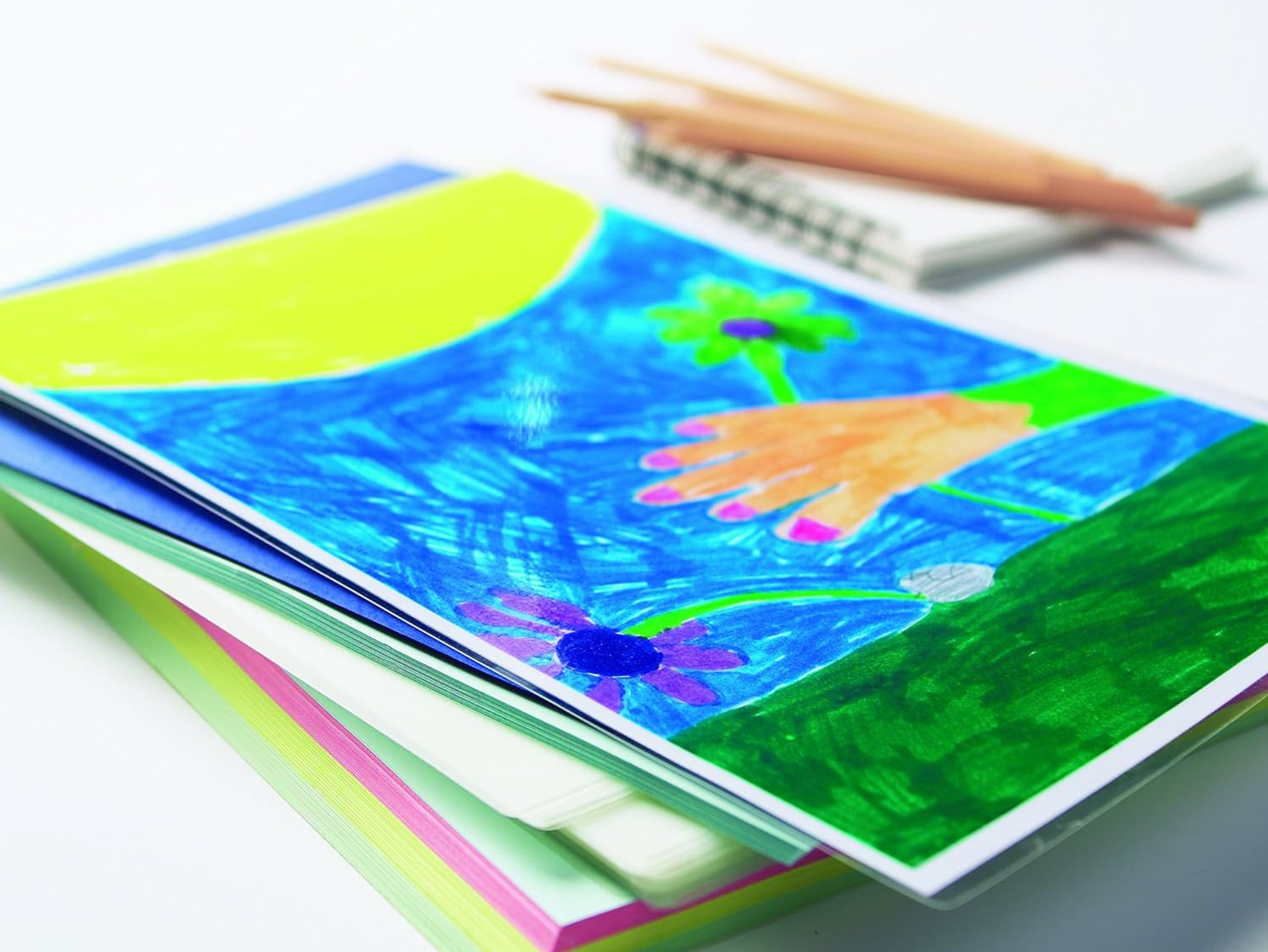 Stapel Zeichnungen mit handgezeichnetem Bild eines Kindes, Buntstifte und Notizblock auf weißem Tisch.