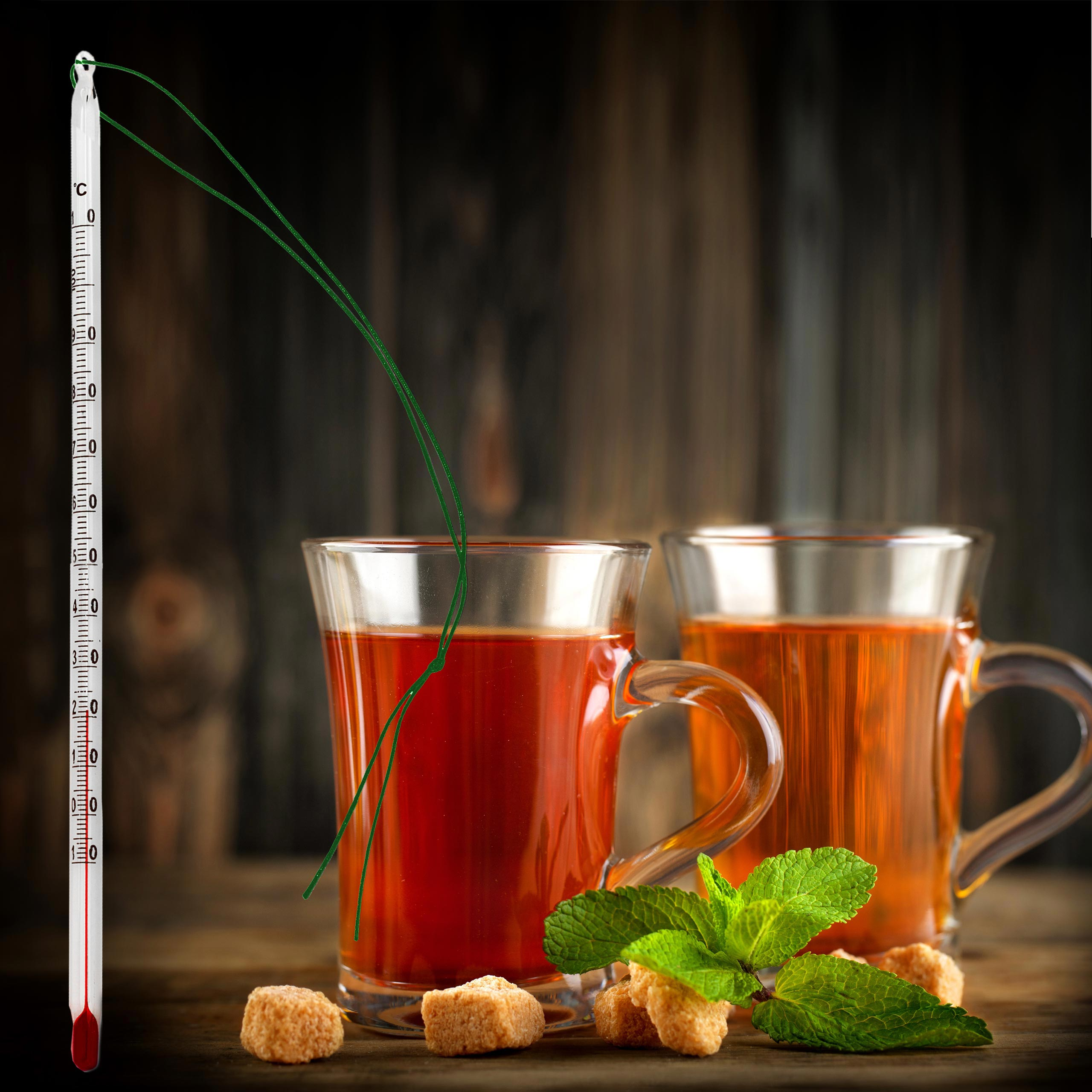 Zwei Teetassen, ein Thermometer, Minzblätter und Würfelzucker auf Holz.