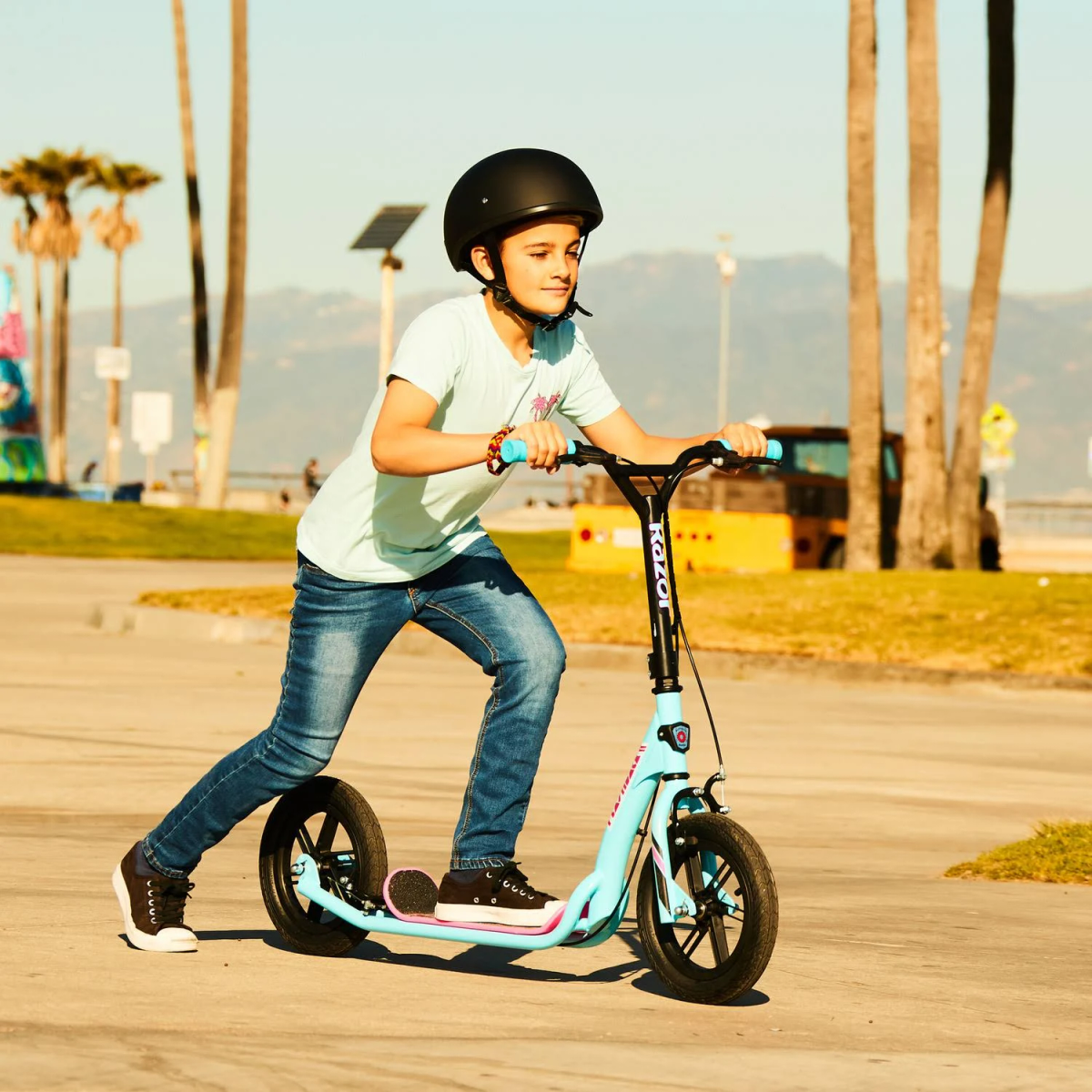 Chłopiec na niebieskiej hulajnodze z czarnym kaskiem, jadący po chodniku. Tło: drzewa i budynki.