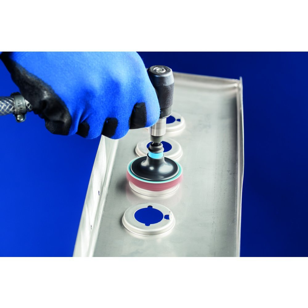 Eine behandschuhte Hand, die einen Schleifer auf einer Metalloberfläche mit kreisförmigen Aussparungen verwendet, blauer Hintergrund.