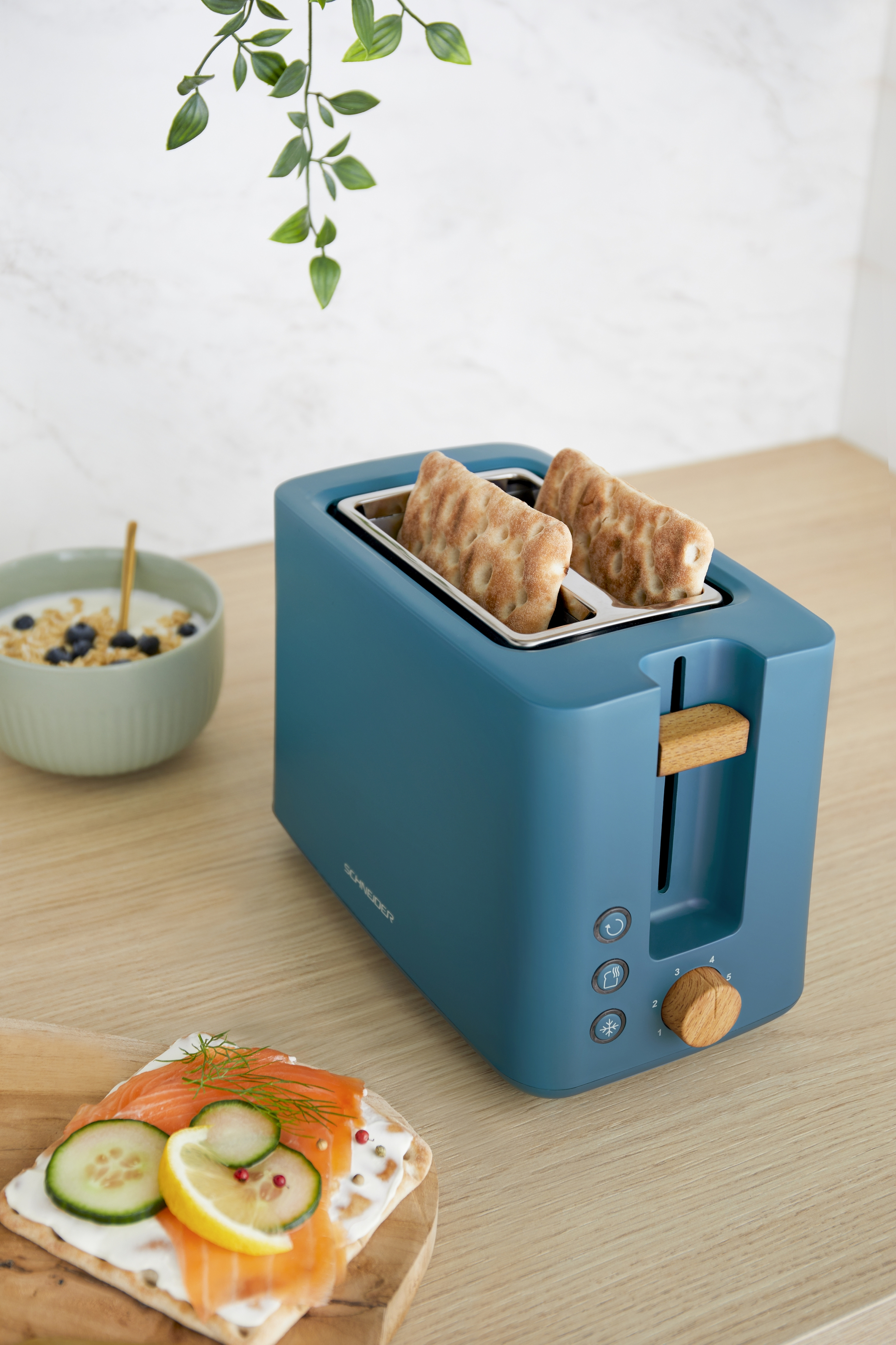 Ein blauer Toaster mit Brot darin. Es gibt einen Holzgriff und Holzknöpfe. Daneben ein Frühstücksteller.