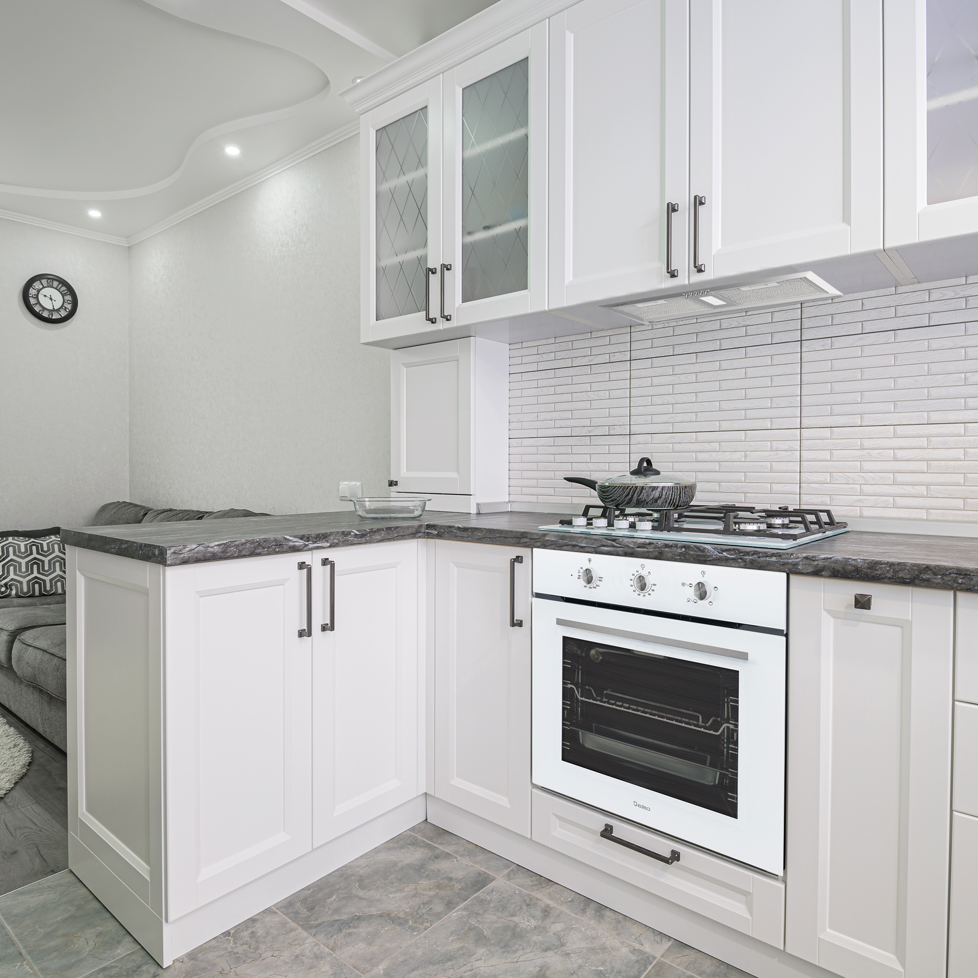 Cucina bianca moderna con mobili, forno e piano cottura, con orologio e divano visibili.