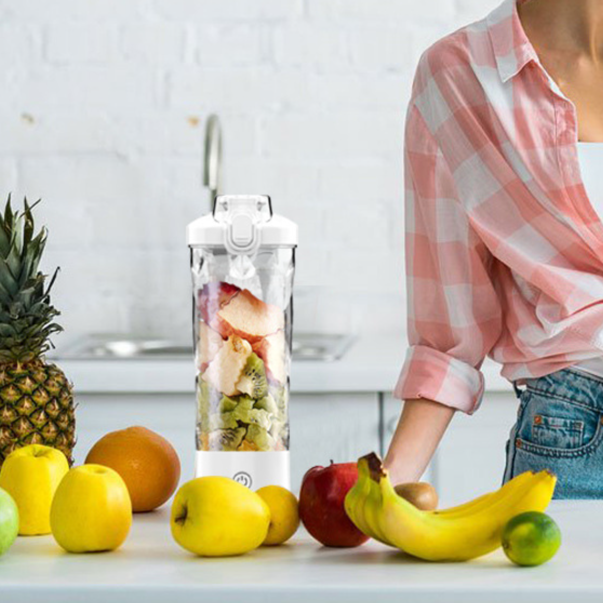 Frau mit einem Mixer, der mit Früchten gefüllt ist. Verschiedene Früchte liegen auf der Theke, wie Äpfel und Bananen.