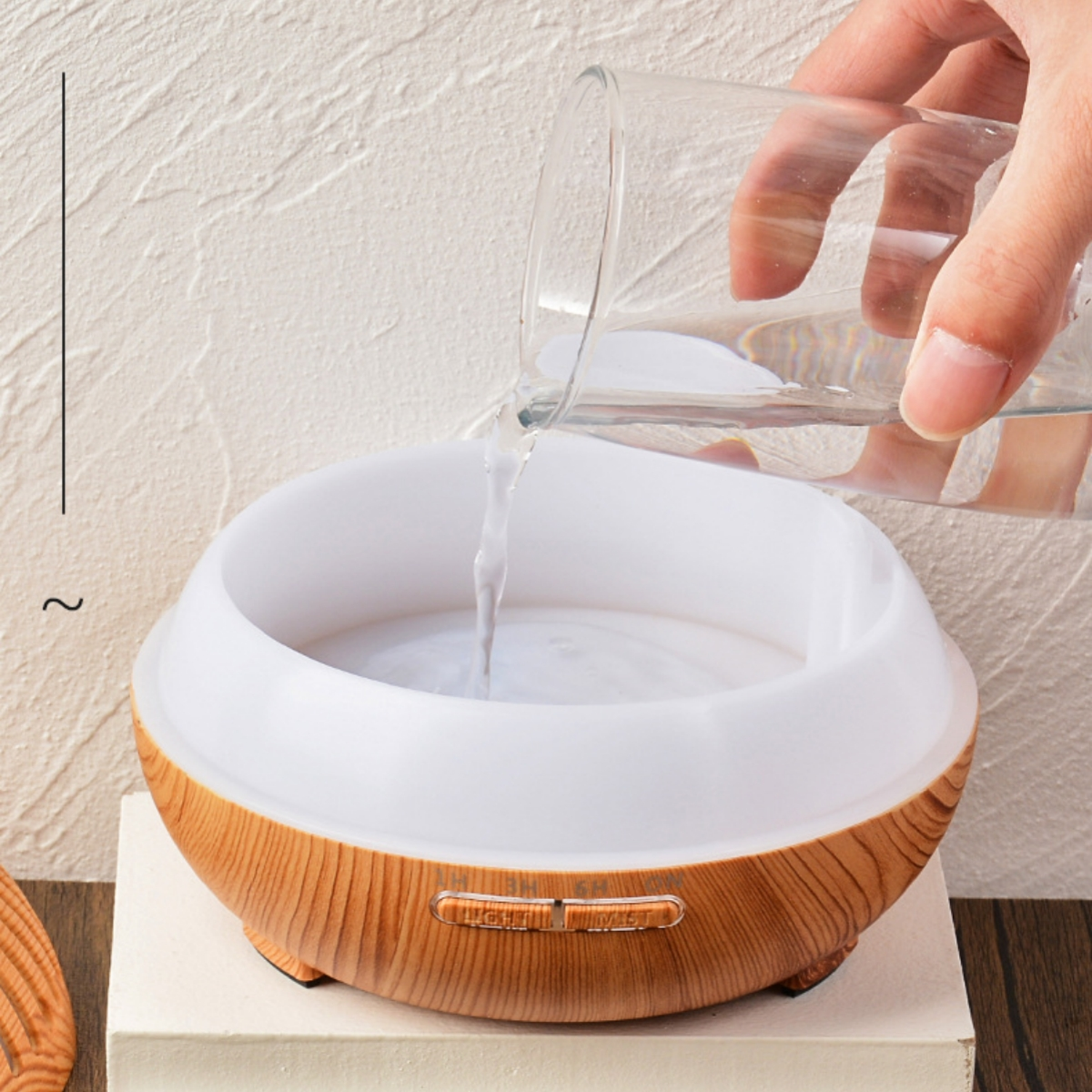 Eine Hand gießt Wasser in einen weißen und holzfarbenen Luftbefeuchter. Text auf dem Luftbefeuchter: Licht, Nebel.