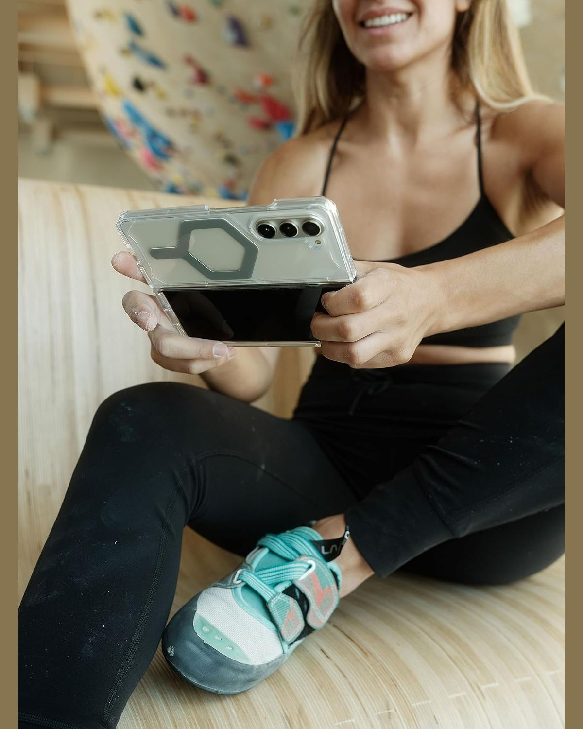 Een vrouw zit met een telefoon in een doorzichtige hoes. Ze draagt sportkleding en klimschoenen.