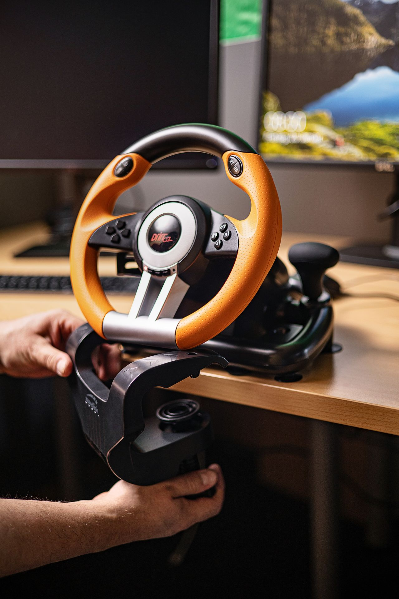 Gaming steering wheel and pedals on a wooden table. The wheel is orange and black.