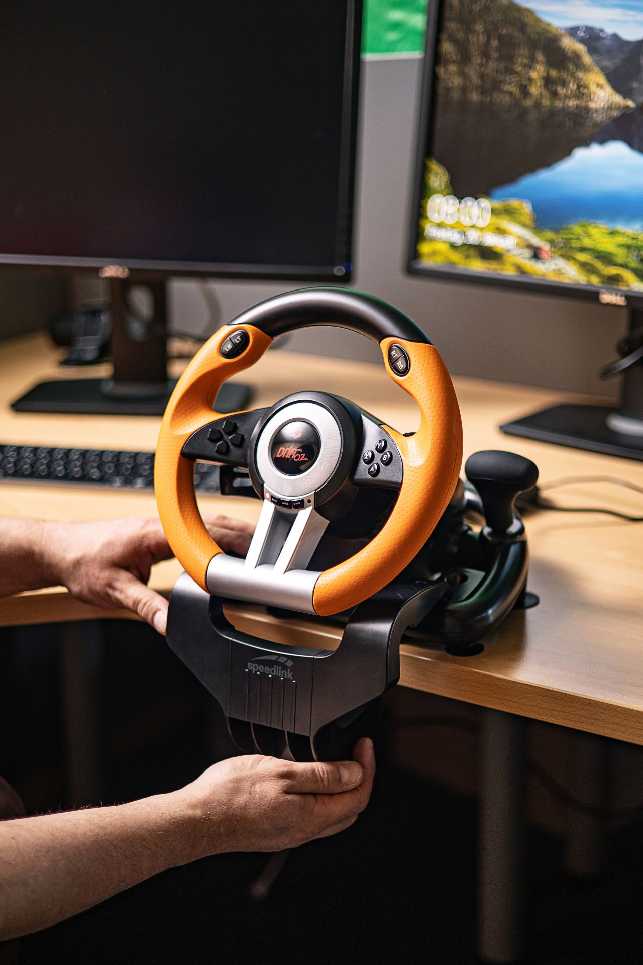 A gaming steering wheel on a desk. Orange and black wheel, with a gear shift lever.