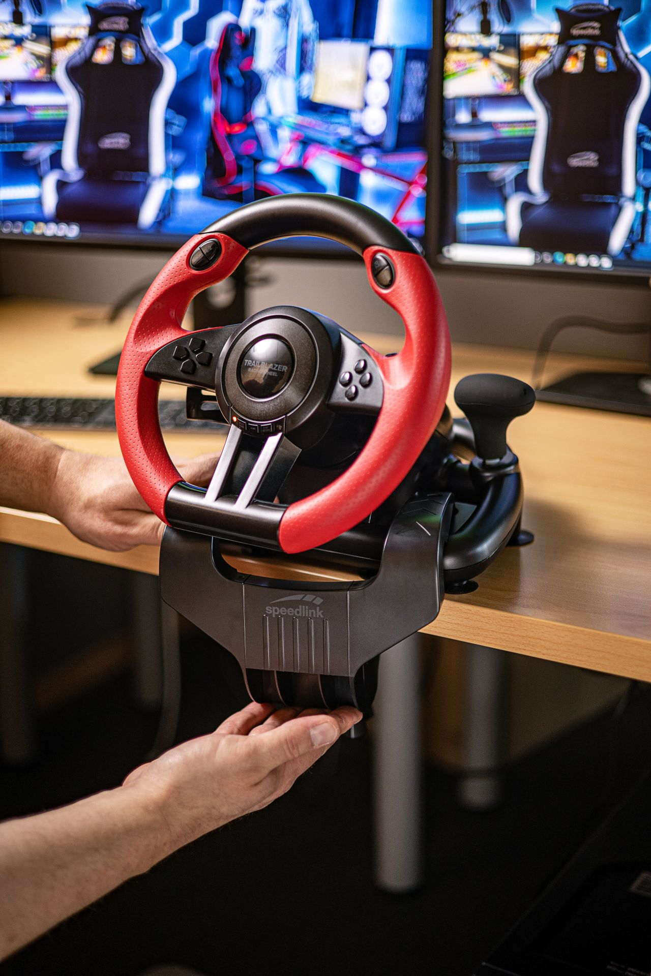 A racing wheel with red accents. Hands are holding the wheel, wood desk in background.