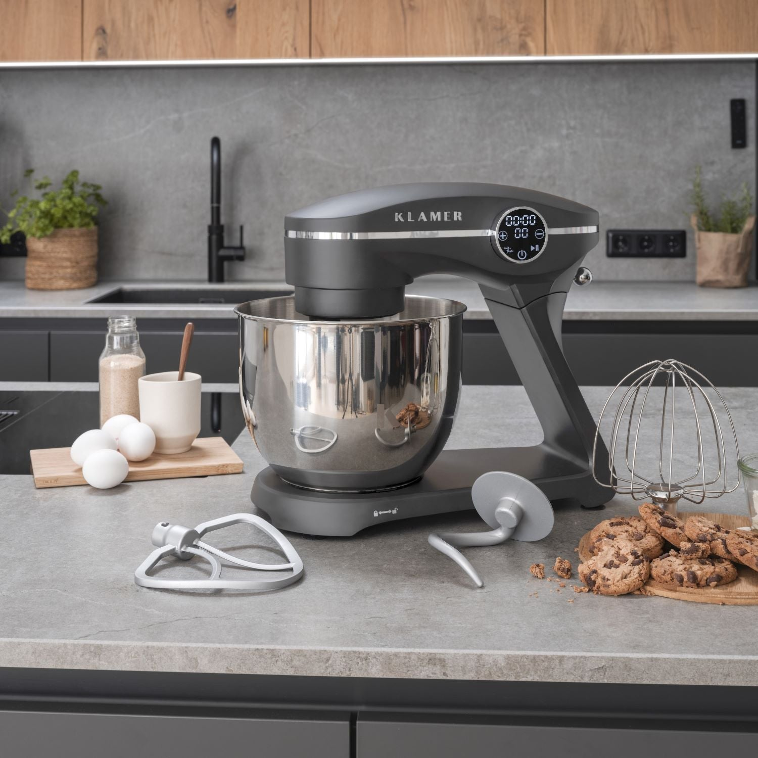 Grauer Küchenmixer mit Edelstahlschüssel. Eier, Kekse und andere Zutaten sind sichtbar.