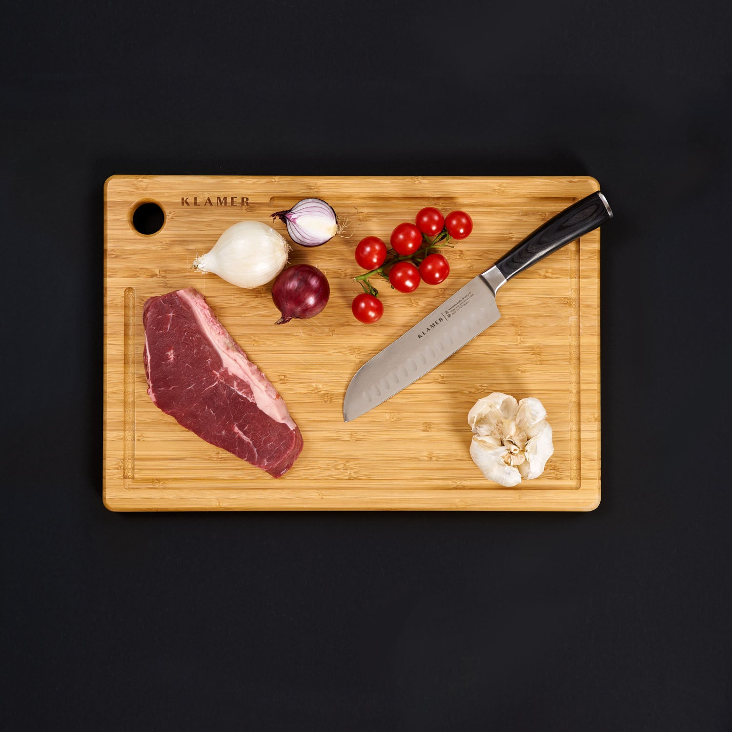 Schneidebrett mit Steak, Tomaten, Zwiebel, Knoblauch und Messer auf schwarzem Hintergrund.