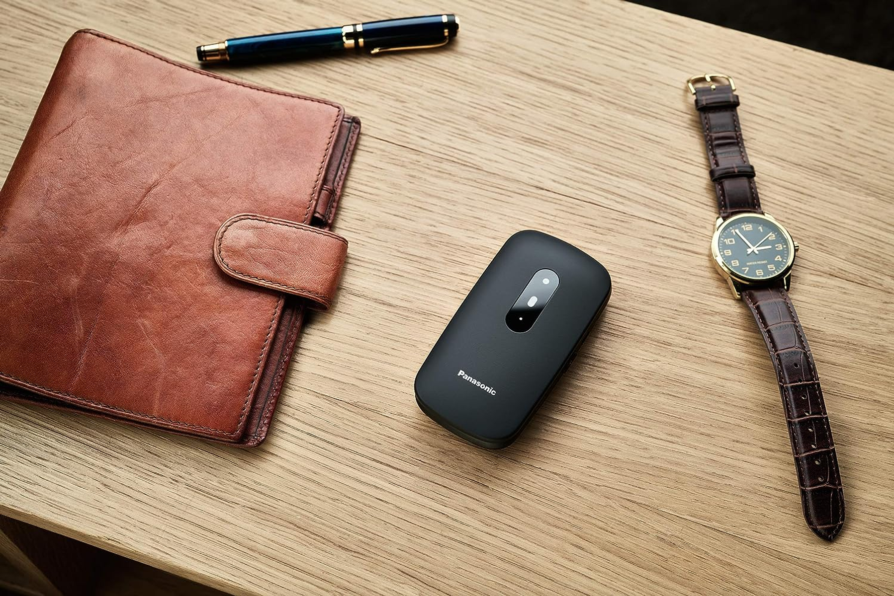 Brązowy skórzany portfel, czarne urządzenie Panasonic, zegarek na rękę i długopis na drewnianej powierzchni.