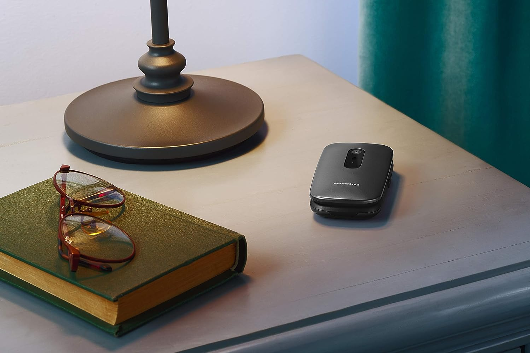 Szafka nocna z lampą, okularami na książce i telefonem z klapką Panasonic. Zielona zasłona w tle.