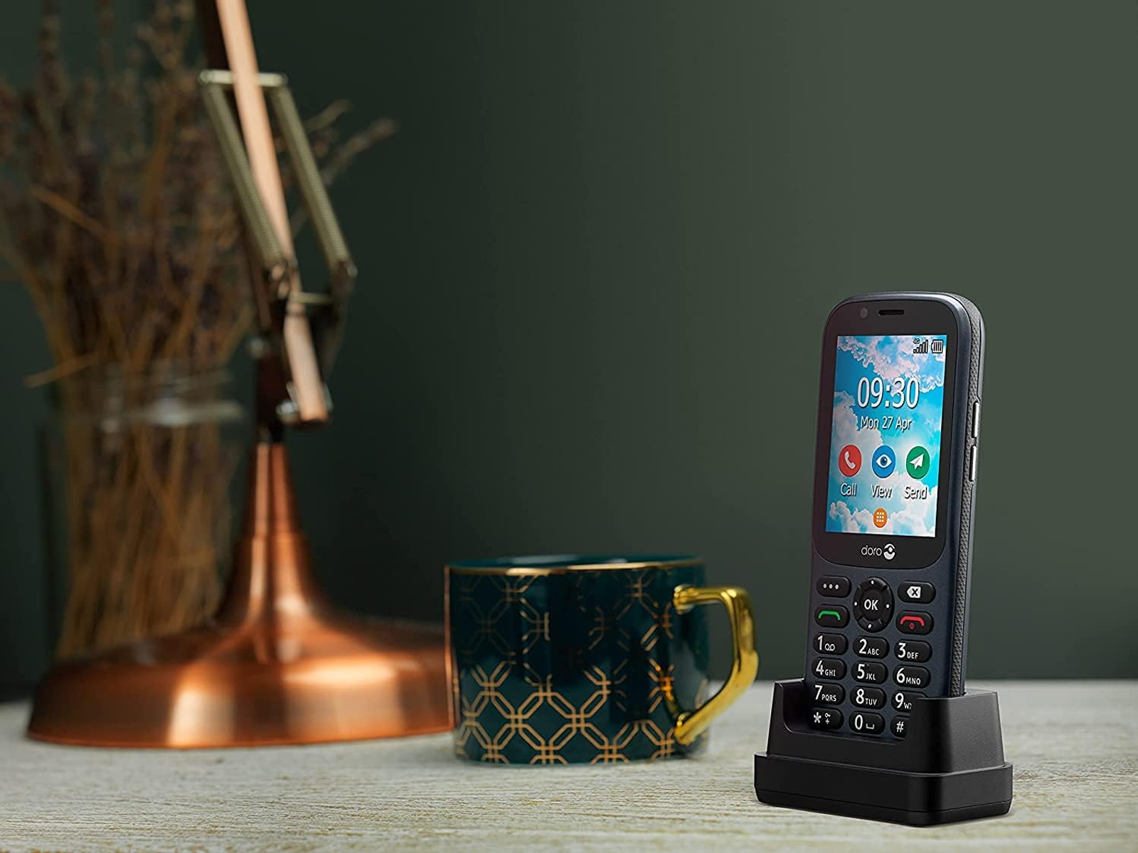 Teléfono Doro en base de carga negra, sobre mesa de madera. Lámpara y taza en el fondo.