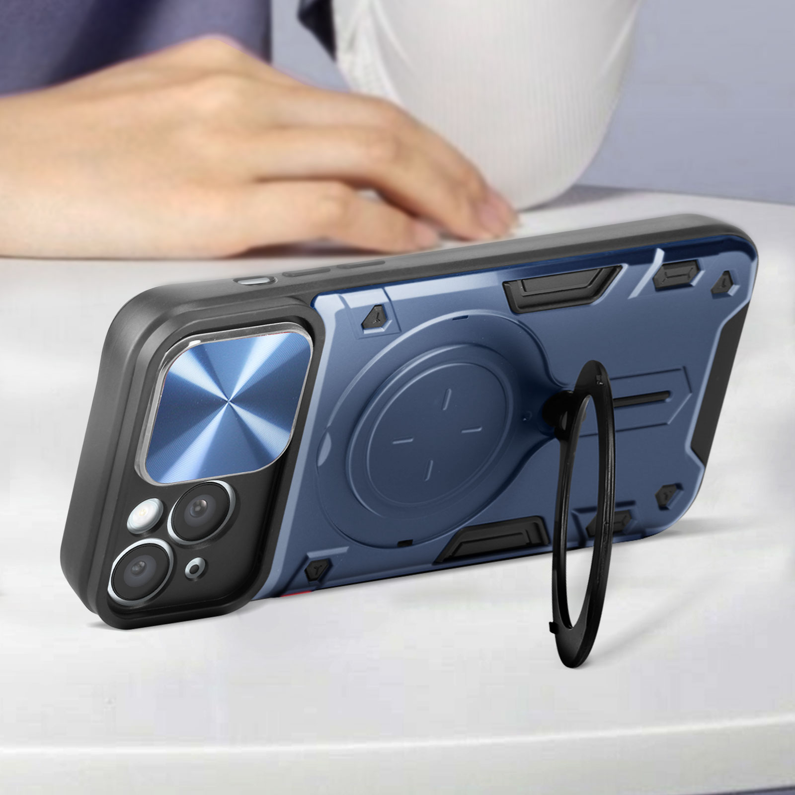 Un cellulare in una custodia blu con supporto ad anello è su un tavolo. Una mano sullo sfondo.