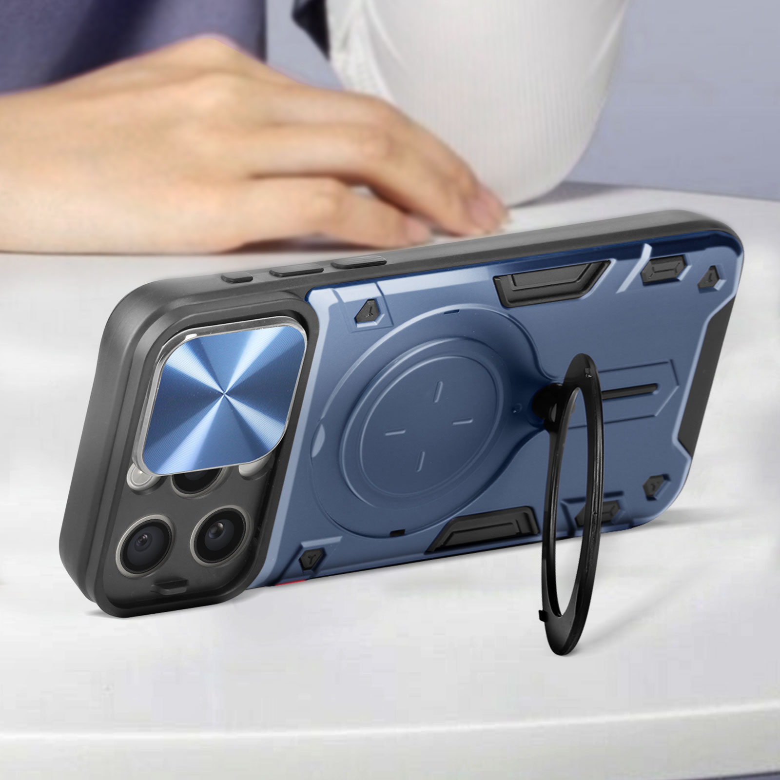 Eine blau-schwarze Handyhülle mit Ringhalterung liegt auf einer weißen Oberfläche. Im Hintergrund eine Hand.