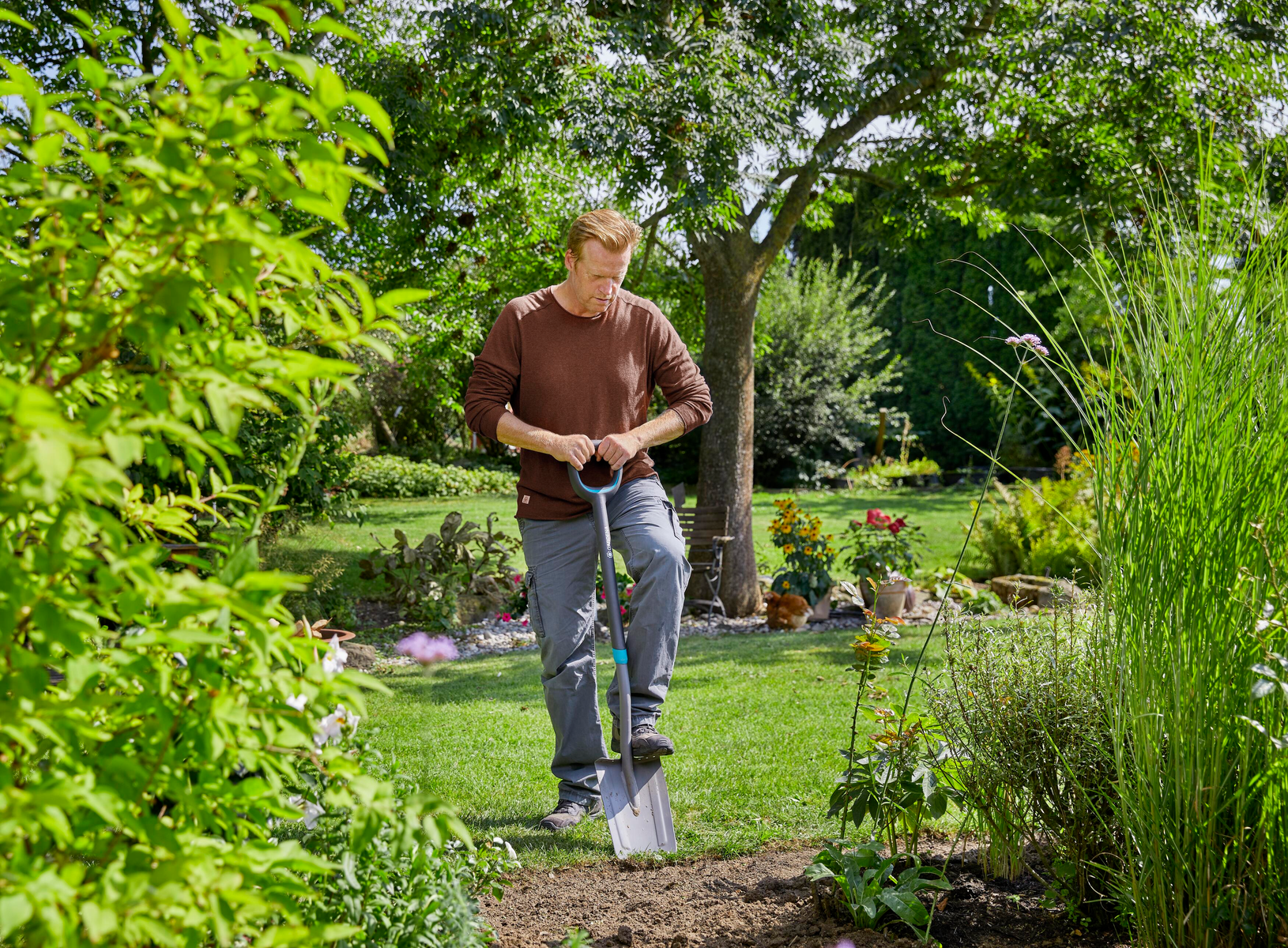 Mann in braunem Hemd benutzt eine Schaufel im Garten. Grünes Gras und Pflanzen um ihn herum.