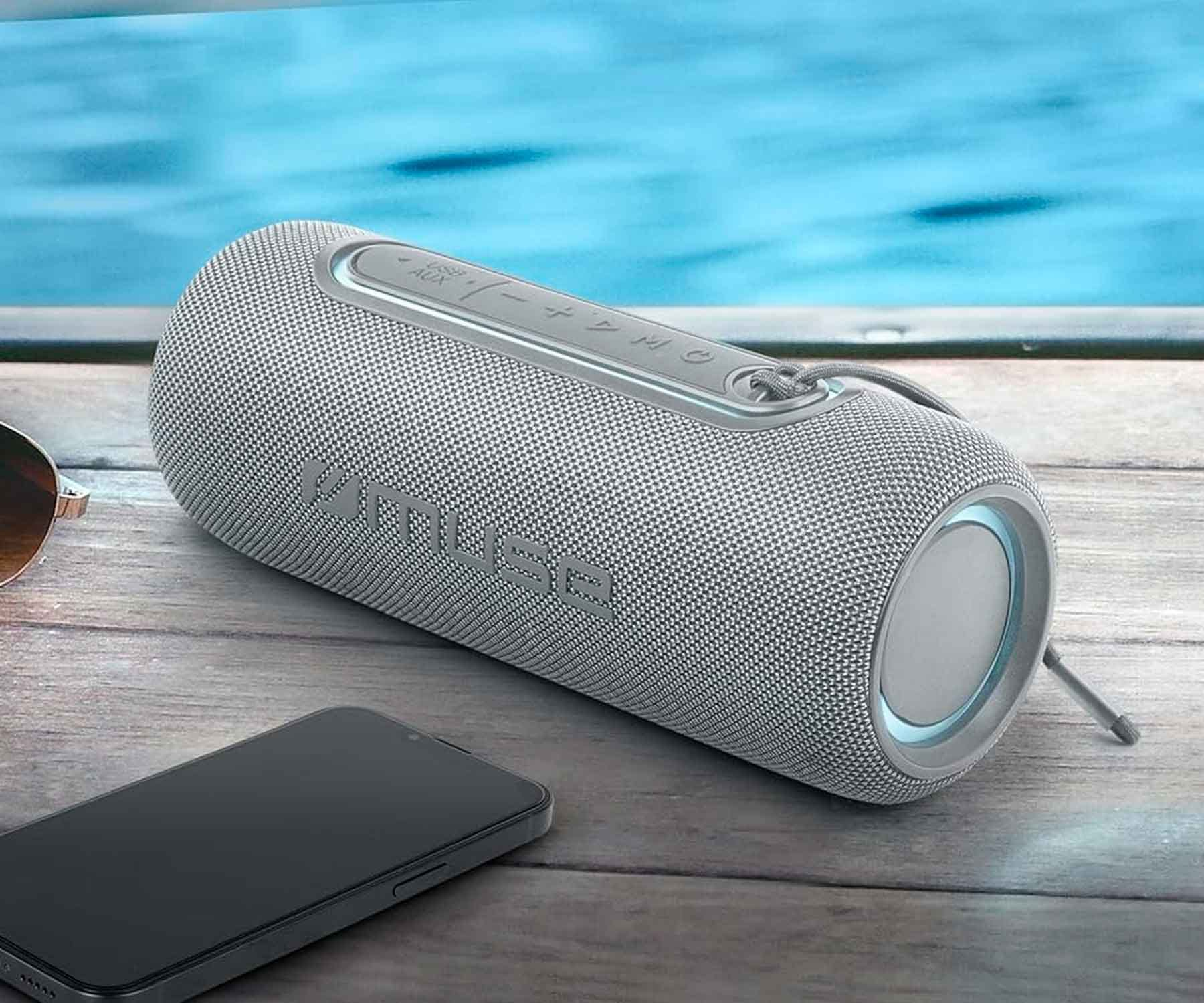 Altoparlante portatile grigio con pulsanti e telefono su superficie di legno vicino all'acqua.