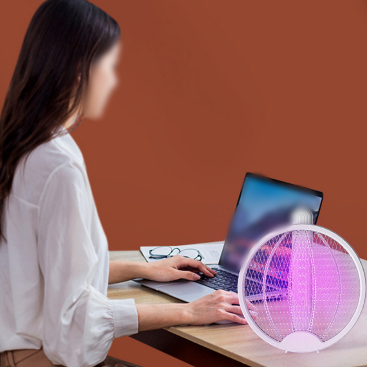 Eine Frau benutzt einen Laptop. Ein Insektenvernichter steht rechts neben ihr. Eine orangefarbene Wand ist im Hintergrund.