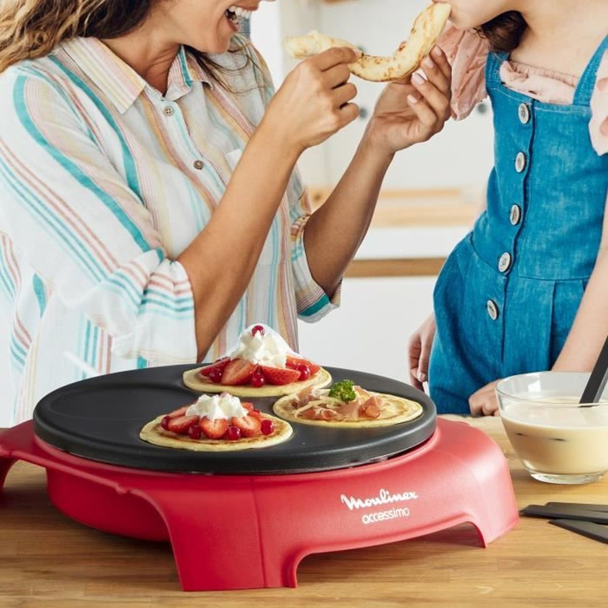 Kobieta karmi dziecko naleśnikiem. Na blacie stoi naleśnikarka z naleśnikami.