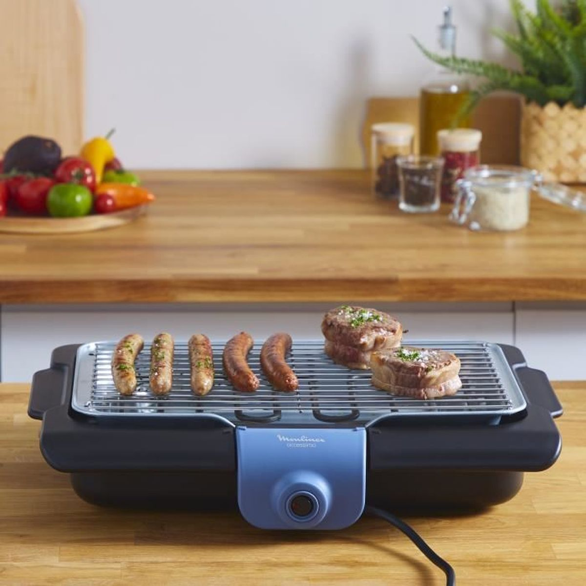 Ein Elektrogrill mit Würstchen und Steaks, Gemüse auf einer Holztheke und Küchenutensilien.