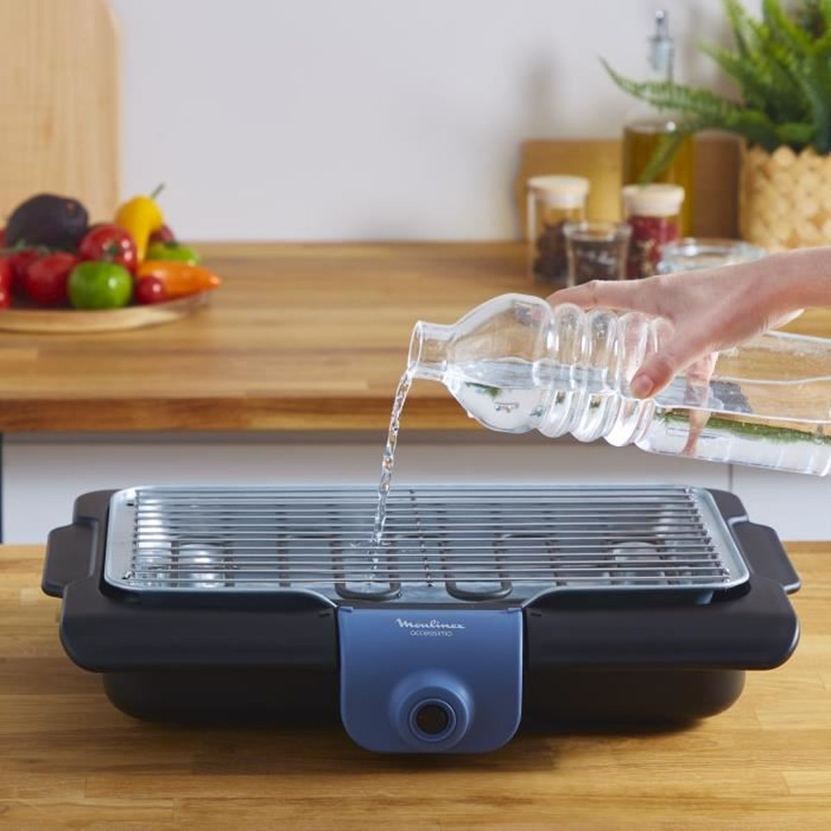 Eine Person gießt Wasser in einen schwarz-blauen Moulinex-Grill auf einer Holz-Arbeitsplatte.