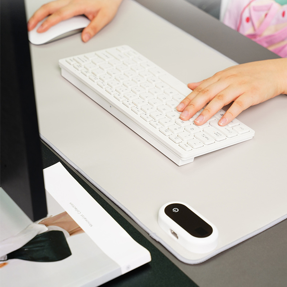 Person tippt auf weißer Tastatur, Maus auf grauer Schreibtischunterlage, Papiere, Monitor sichtbar.