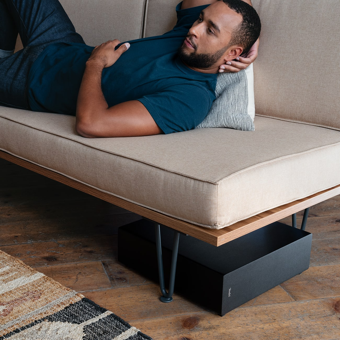 Mann auf beigem Sofa mit schwarzer Box darunter. Holzboden, Teppich unten links.