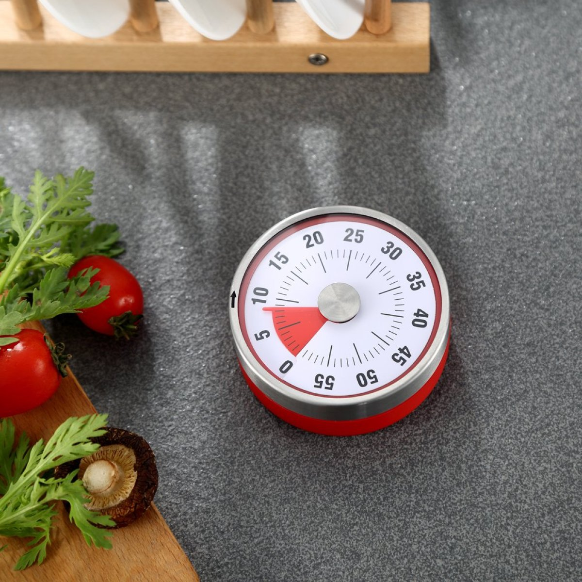 Roter und silberner Timer auf grauer Oberfläche. Tomaten, Petersilie und ein Pilz liegen daneben.